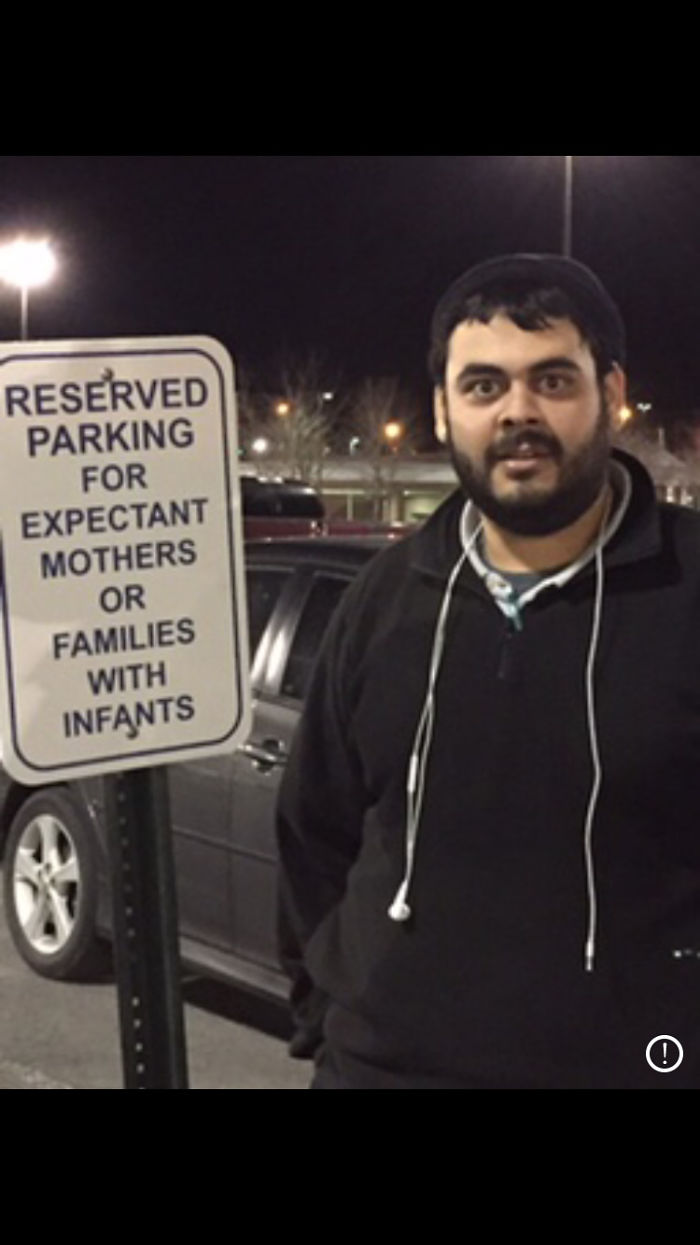 Man standing humorously by an expectant mothers parking sign, with earbuds hanging from a black shirt.