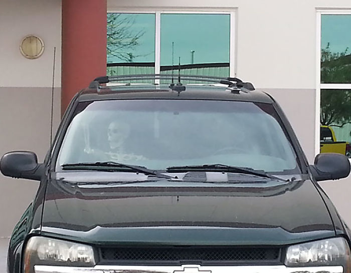 A skeleton, humorously posed as a driver, is seen inside a parked SUV.