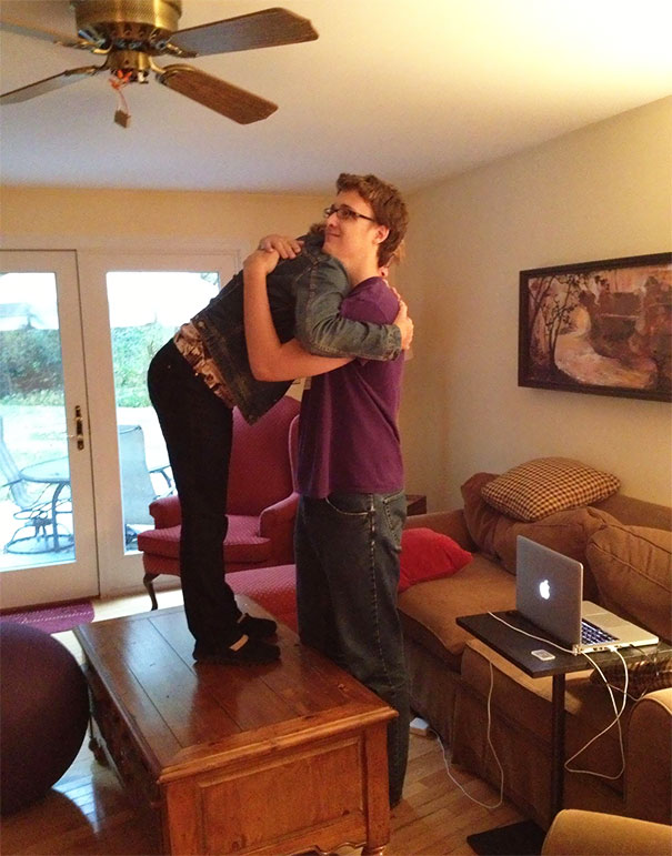 Tall person standing on a table hugging another tall person in a living room, showcasing everyday struggles of tall people.