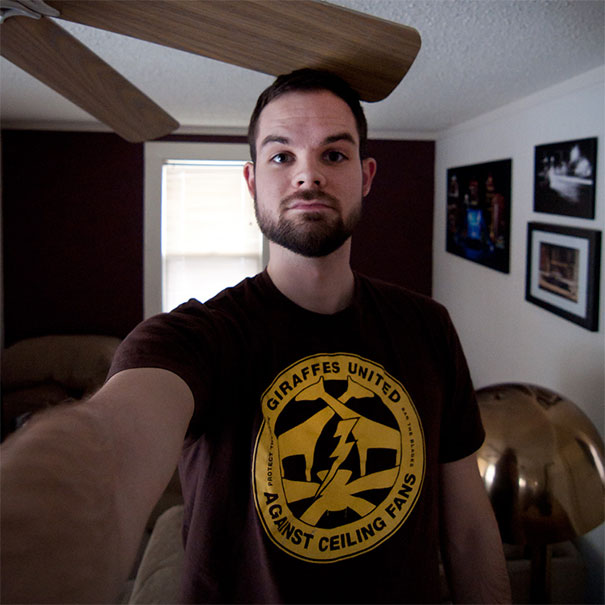 Tall person taking selfie with a ceiling fan close to his head, wearing a shirt about struggles with low ceilings.