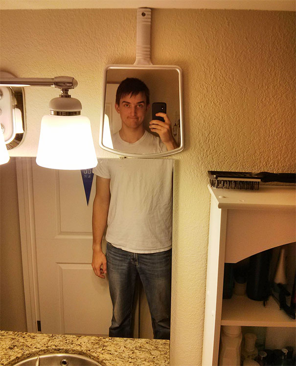 Man taking mirror selfie using a small wall mirror above bathroom counter, illustrating everyday struggles of tall people.