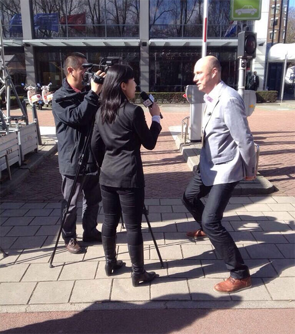 Tall man leaning to fit in camera frame during an outdoor interview, highlighting everyday struggles of tall people.
