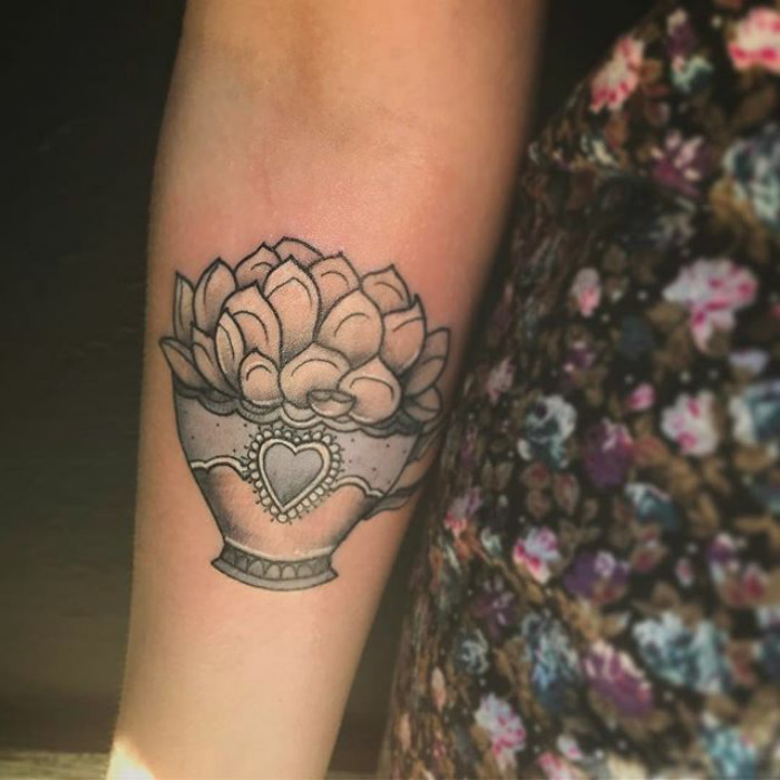 Succulent tattoo on forearm featuring potted plant design with heart detail in black and grey ink.