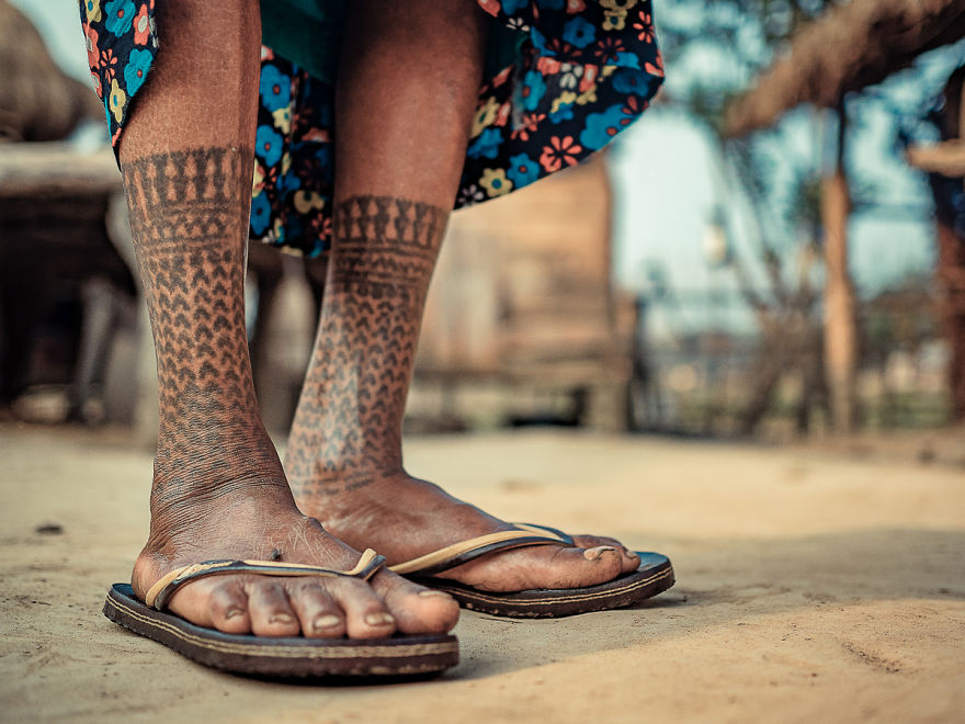 Close-up of tattooed legs of a Tharu tribe woman, wearing flip-flops on a sandy surface.