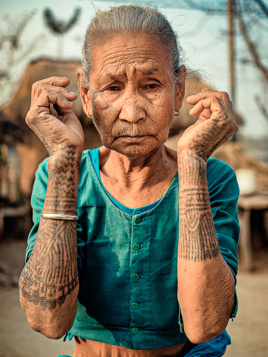 Elderly Tharu woman with traditional tattoos on her arms and wearing a teal blouse.