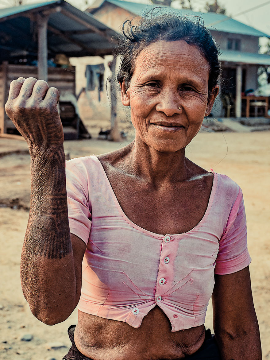Tharu tribe woman displaying traditional tattoos with a smile, wearing a pink top in a village setting.