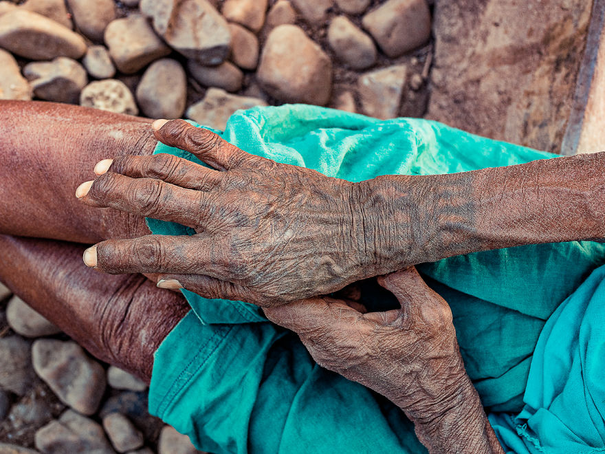 Tattooed hands of an elderly Tharu woman, resting on a turquoise cloth background with stone pebbles nearby.