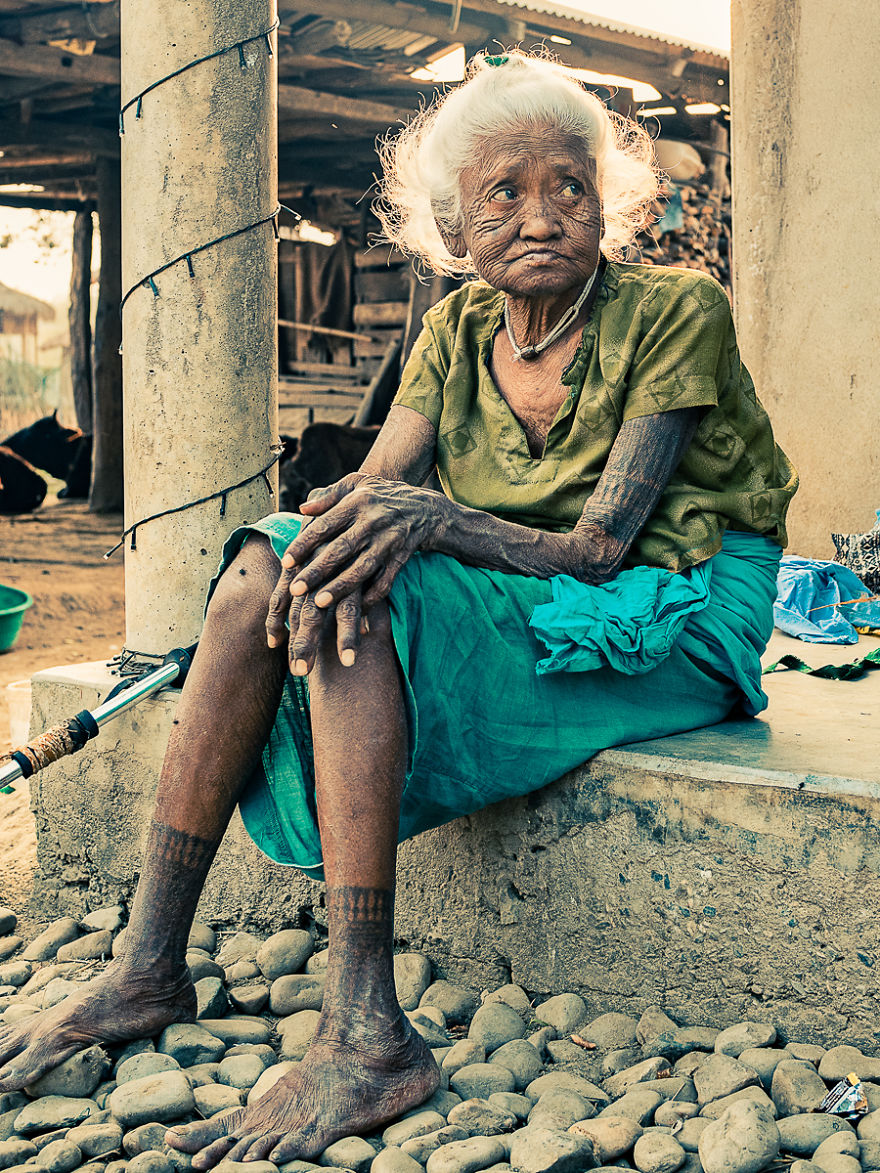 Elderly Tharu woman with traditional tattoos, sitting on a stone step in a rural setting.