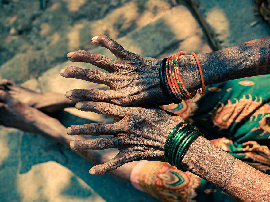 Elderly Tharu woman's tattooed hands with colorful bangles, seated outdoors.