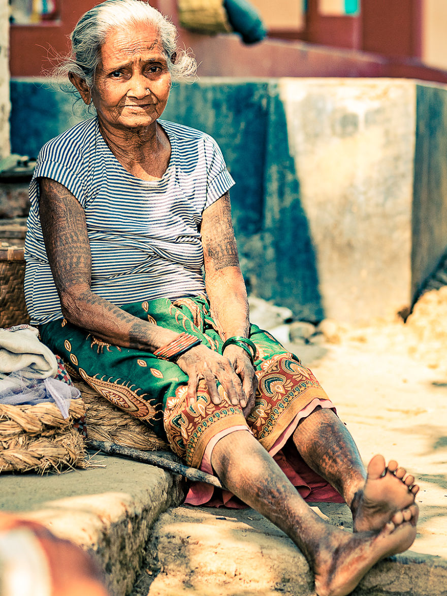 Elderly woman from the Tharu tribe with traditional tattoos, sitting outdoors in colorful clothing.
