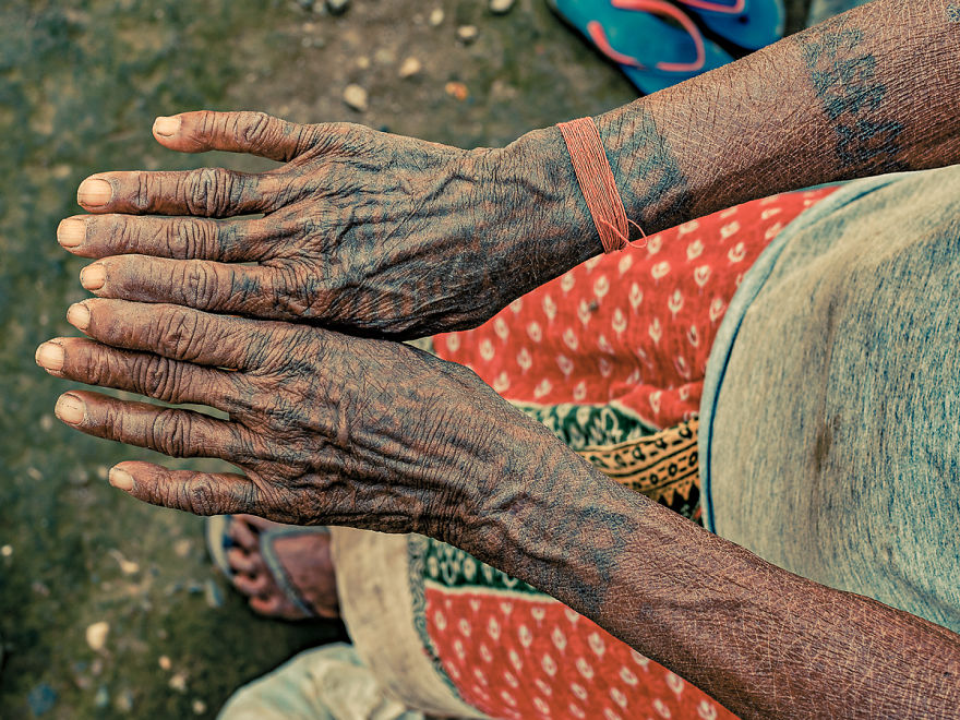 Tattooed hands of an elderly Tharu woman, showcasing traditional body art against a colorful skirt background.