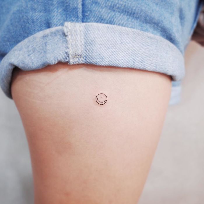 Minimalist tiny smiley face tattoo on inner thigh, under rolled-up denim shorts.