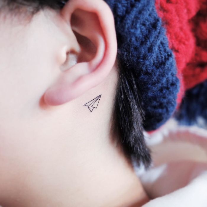 Minimalist paper airplane tattoo behind ear, showcasing discreet tattoo artistry.