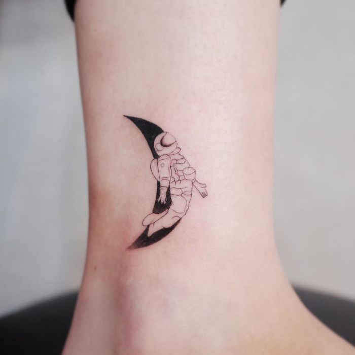 Minimalist tattoo of an astronaut floating on a crescent moon on skin.