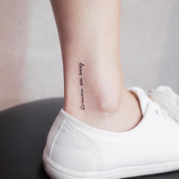 Minimalist tattoo on ankle reading "love me more" with a heart, showcased with white shoes for discreet style.