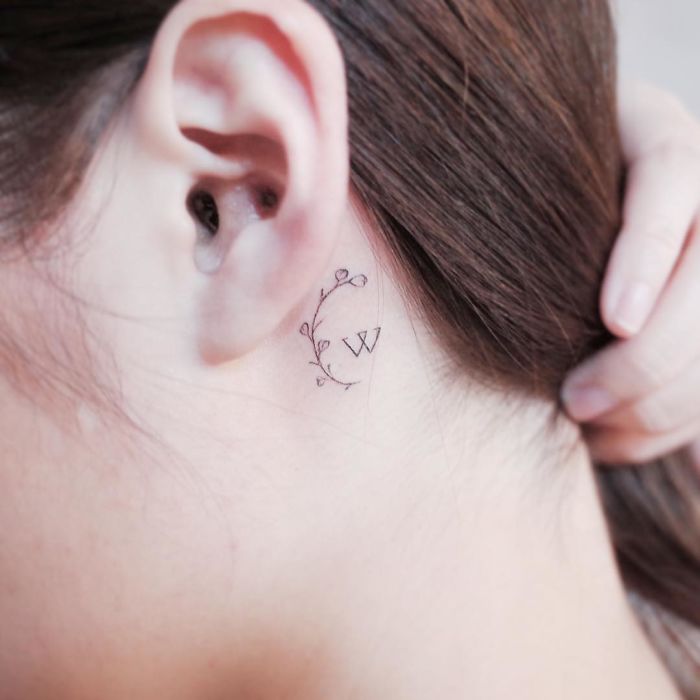 Minimalist tiny tattoo of a floral vine and letter "W" behind a person's ear.