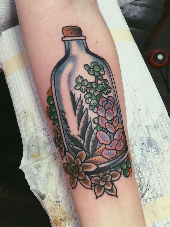 Colorful succulent tattoo in a bottle design on forearm, featuring various vibrant succulents and floral elements.