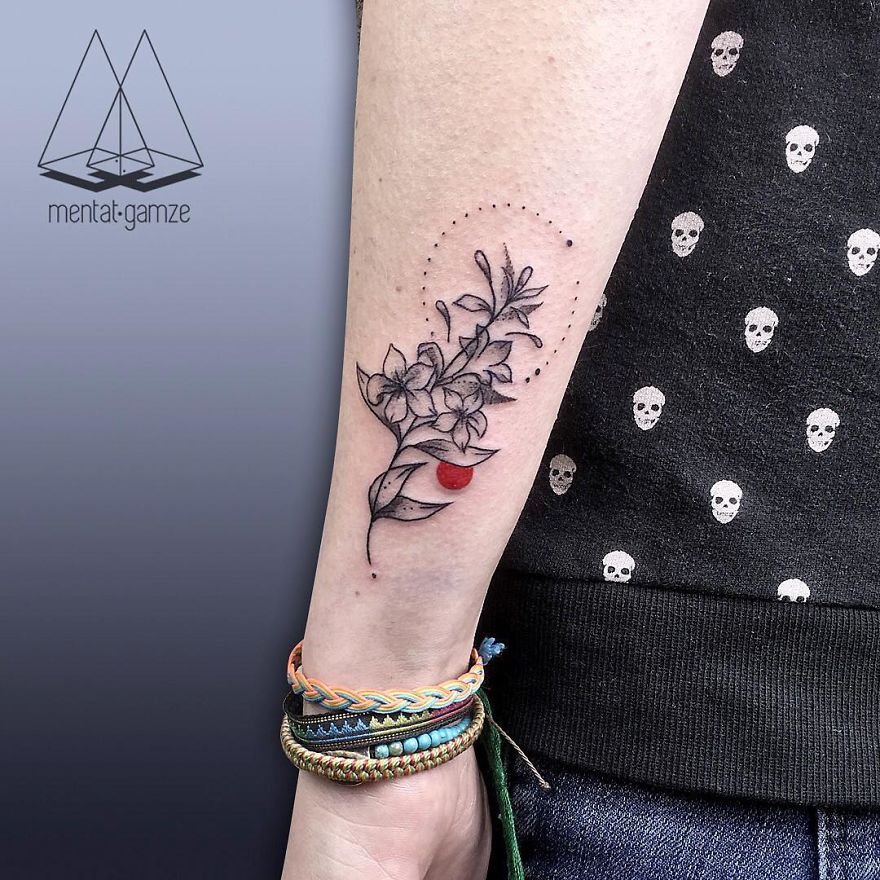 Tattoo artist designs floral and red dot tattoo on forearm, wearing skull-patterned shirt and colorful bracelets.