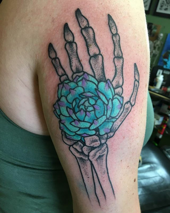 Succulent tattoo featuring a skeleton hand holding a vibrant green and purple plant on an upper arm.