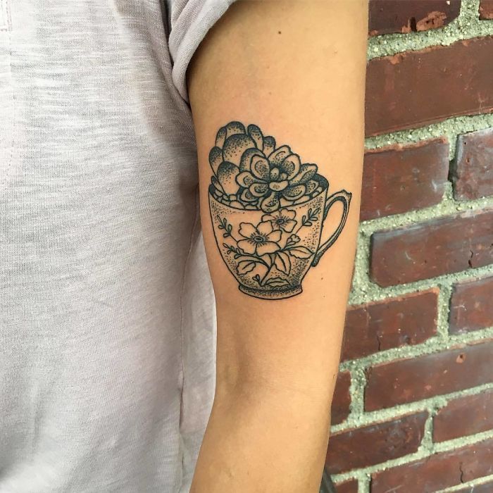 Succulent tattoo in a teacup design on a person's arm, showcasing intricate line art against a brick wall background.
