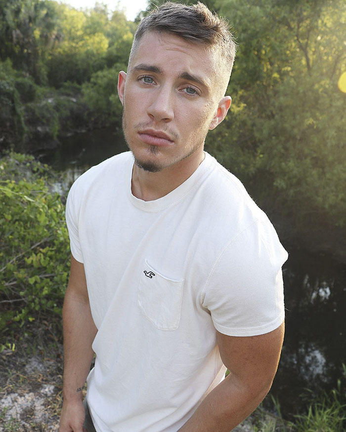 A person standing outdoors, showcasing female-to-male transition, wearing a white shirt with trees and a river in the background. A person standing outdoors, showcasing female-to-male transition, wearing a white shirt with trees and a river in the background.