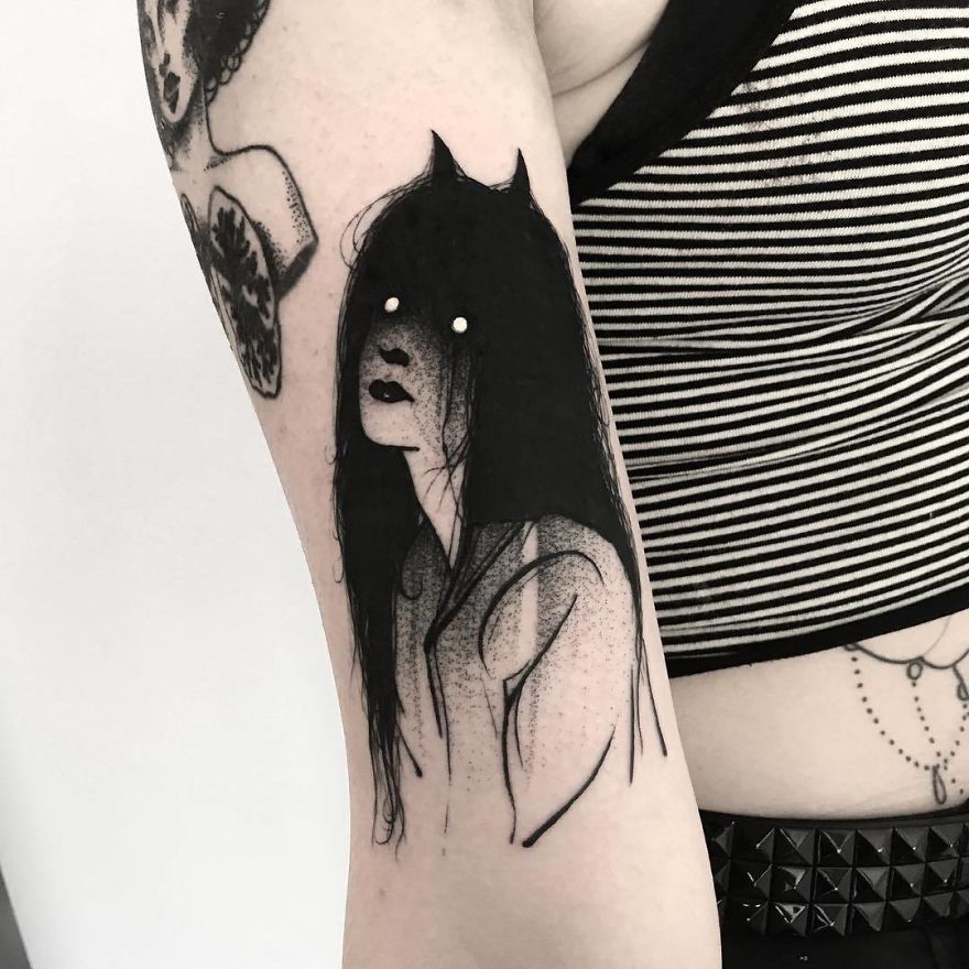 Faceless girl tattoo with dark hair and hollow eyes on upper arm of a person wearing a striped top.