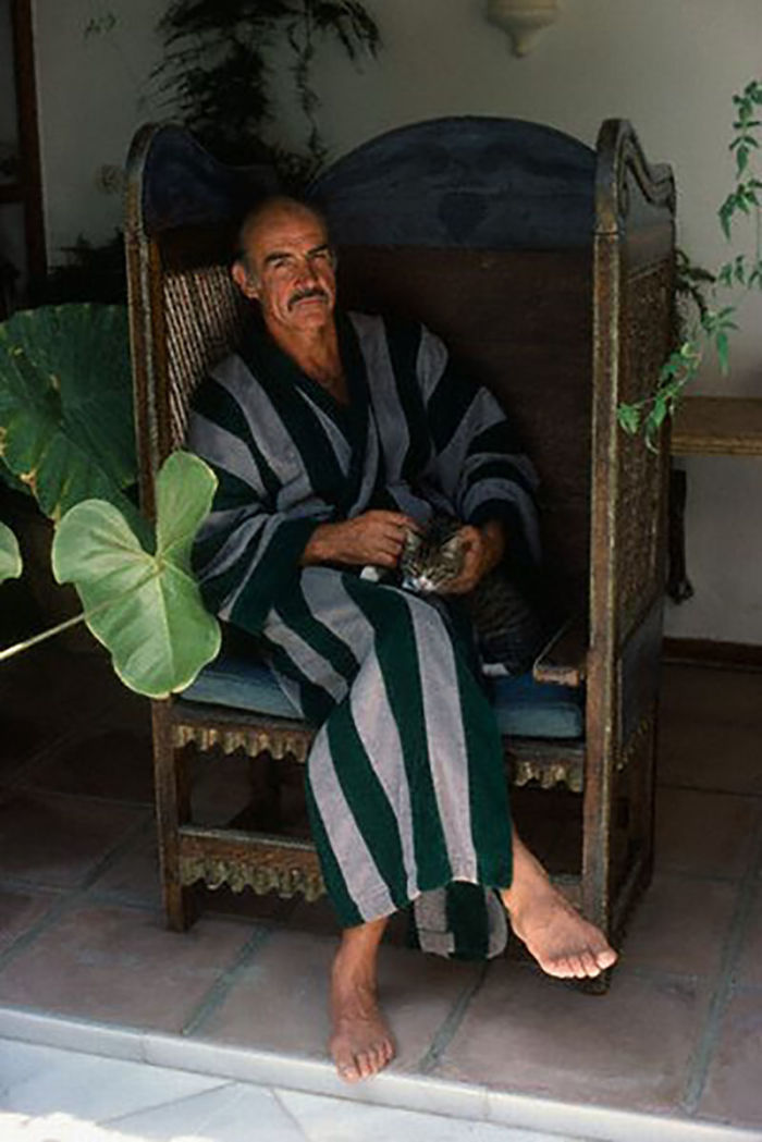 Man in striped robe sitting barefoot on a wooden chair petting a cat surrounded by plants, famous people and their cats.