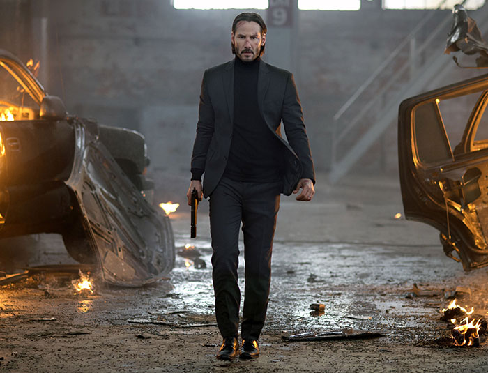 John Wick walking through burning wreckage holding a gun in an action scene from the movie, illustrating fun facts about movies.