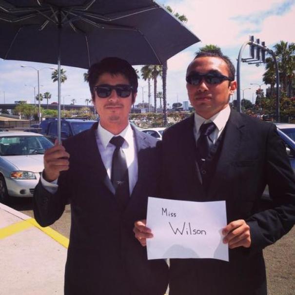 Two men in suits holding a humorous airport pickup sign for "Miss Wilson" at an airport.