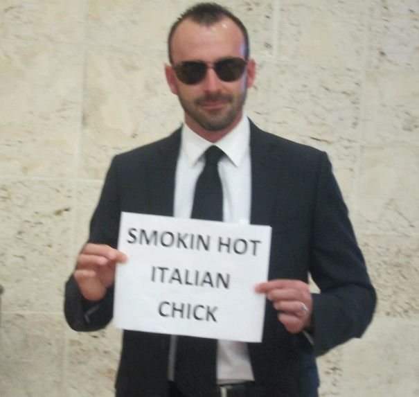 Man in a suit and sunglasses holding a funny airport pickup sign reading "Smokin Hot Italian Chick."