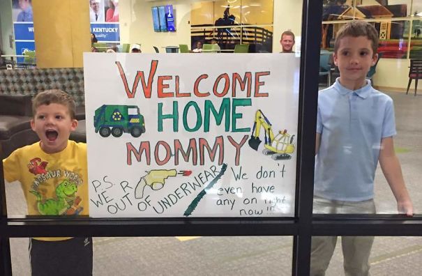 Two kids holding a funny airport pickup sign welcoming their mom, featuring drawings and an amusing message about underwear.