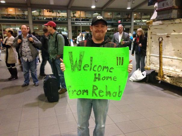 Man holding a funny airport pickup sign saying "Welcome Home From Rehab" in a bustling terminal.