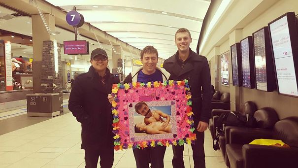 Three men at an airport holding a funny, colorful pickup sign with a large baby photo.