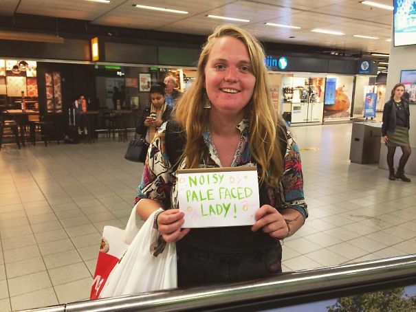 Woman holding a funny airport pickup sign reading "Noisy Pale Faced Lady!" at an airport terminal.