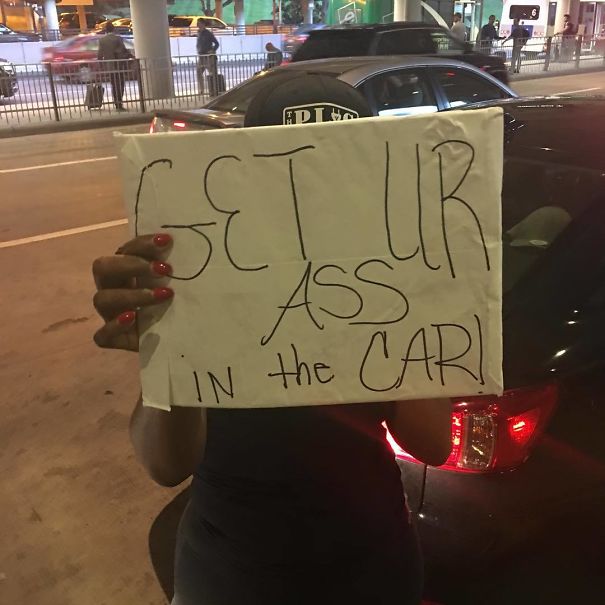 Funny airport pickup sign reading "Get ur a*s in the car" held by a person in a parking area.