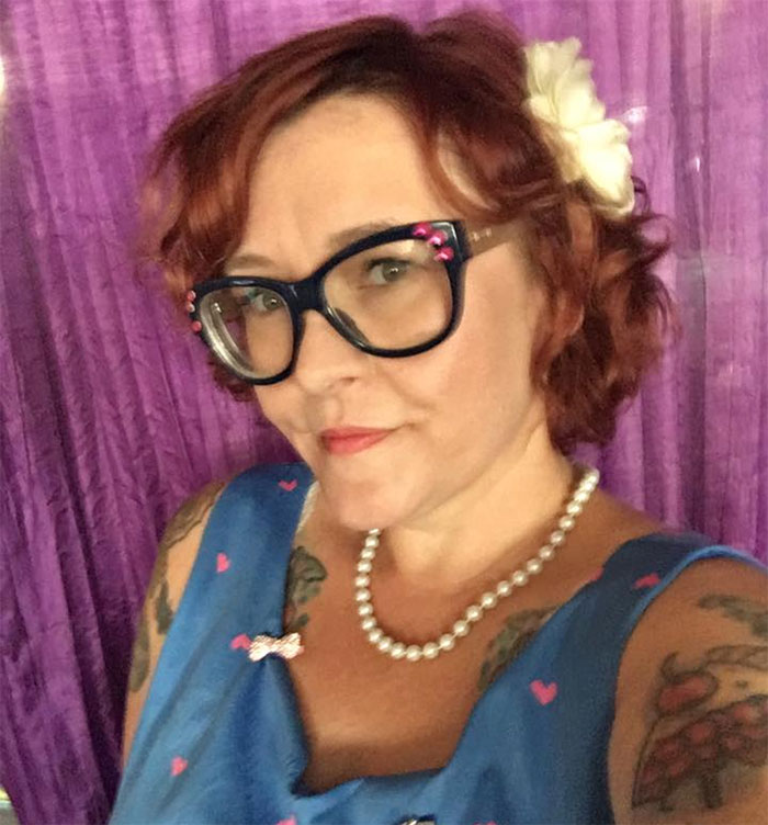 Person with glasses and tattoos, wearing a blue dress and pearls, posing in front of a purple curtain; depression symptoms.