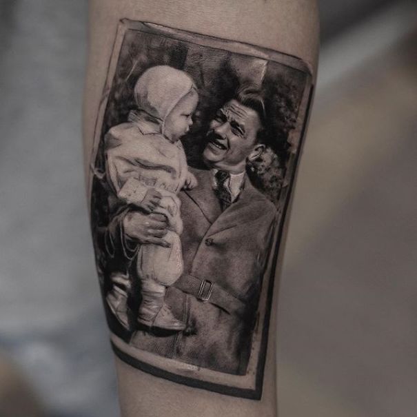 Realistic tattoo of a man holding a child, designed by a Belgian tattooist, resembling a printed photograph on skin.