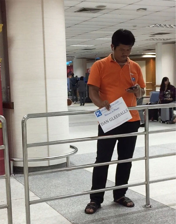 Man in an orange shirt holding a funny airport pickup sign with a humorous name, looking at his phone.