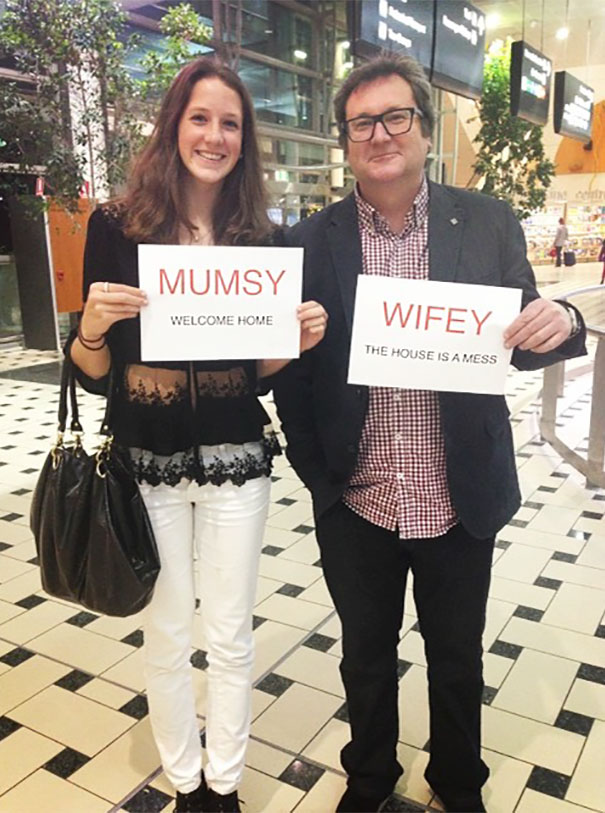 Two people holding funny airport pickup signs: "Mumsy, welcome home" and "Wifey, the house is a mess."