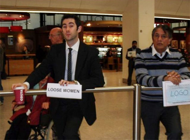 Man in a suit holding a funny airport pickup sign labeled "Loose Women" at an airport terminal.