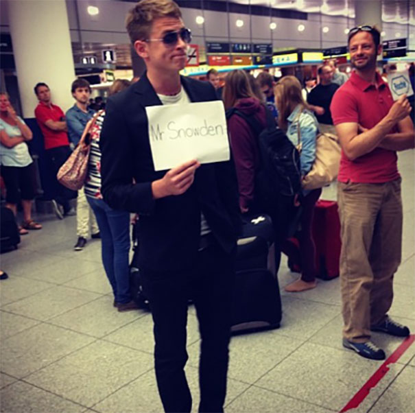 Man holding a funny airport pickup sign reading "Mr Snowden" in a busy terminal.