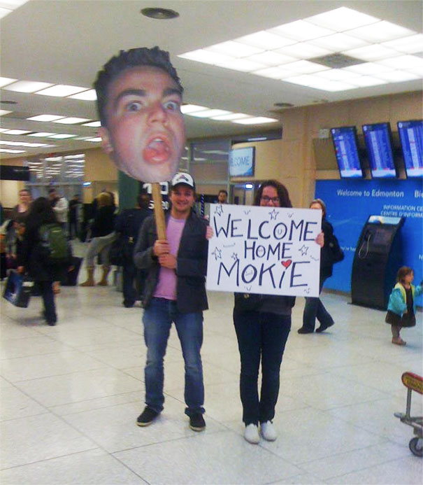 Funny airport pickup sign with giant face cutout and welcome message in a busy terminal.