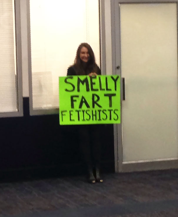 Woman holding a humorous airport pickup sign that reads "Smelly Fart Fetishists" in a terminal.