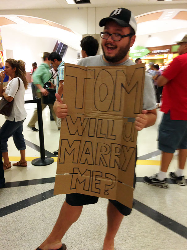 Man at airport holding a funny pickup sign reading "Tom, will you marry me?" causing laughter among bystanders.
