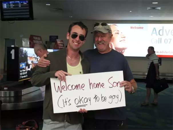 Two people at the airport holding a funny welcome sign saying, "Welcome home son (It's okay to be gay)."