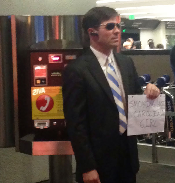 Man in sunglasses holding a funny airport pickup sign near a payphone station.