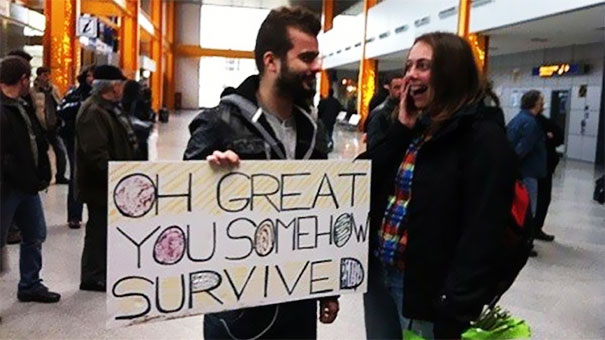 Man holding a funny airport pickup sign reading "Oh great you somehow survived" while making a woman laugh.