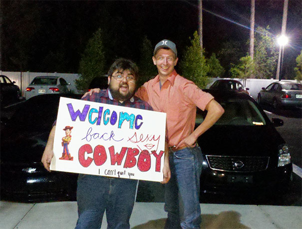 Two men holding a funny airport pickup sign that says "Welcome back s**y cowboy" in a parking lot.