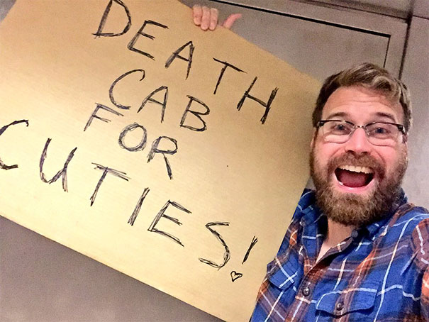 Man holding a funny airport pickup sign saying "Death Cab for Cuties," smiling widely in a plaid shirt.