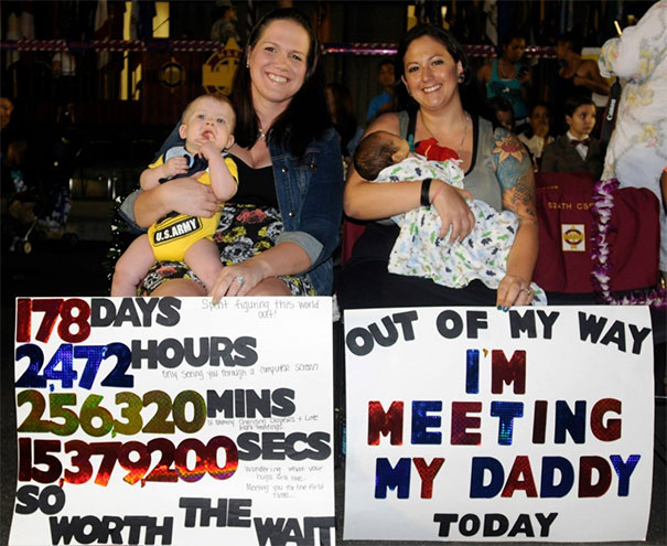 Two women hold funny airport pickup signs with babies on their laps, causing chuckles.